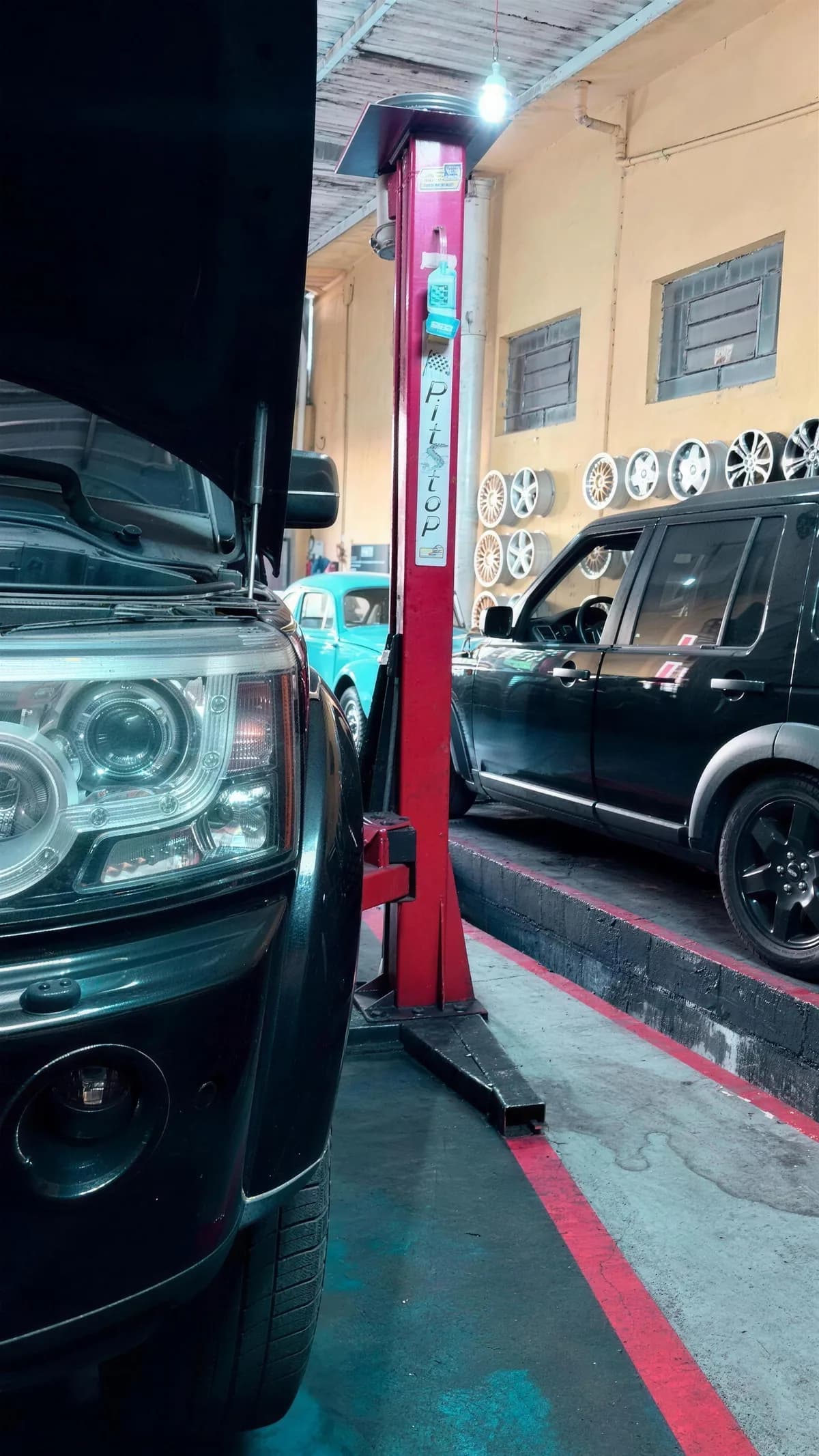 Land Rover preto de capô aberto ao lado do elevador vermelho, com outro Land Rover e um carro azul-claro ao fundo