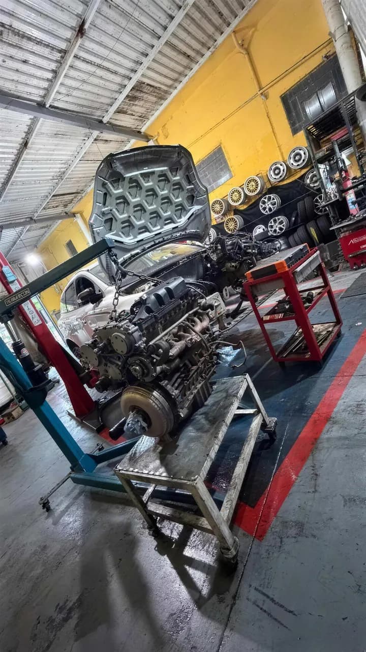 Motor içado pelo guincho ao lado de um carro branco de capô aberto, com a parede de rodas ao fundo