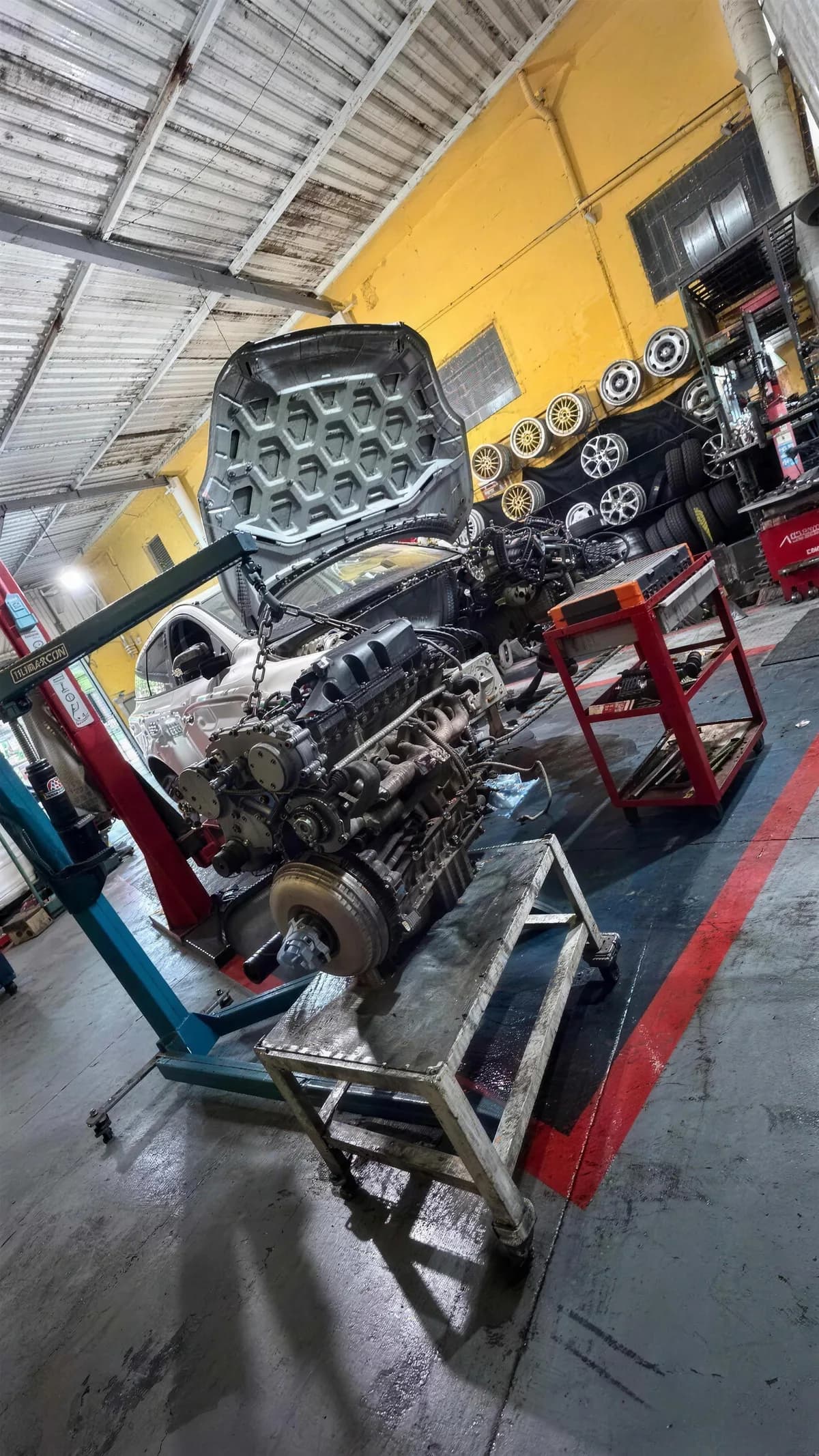 Motor içado pelo guincho ao lado de um carro branco de capô aberto, com a parede de rodas ao fundo