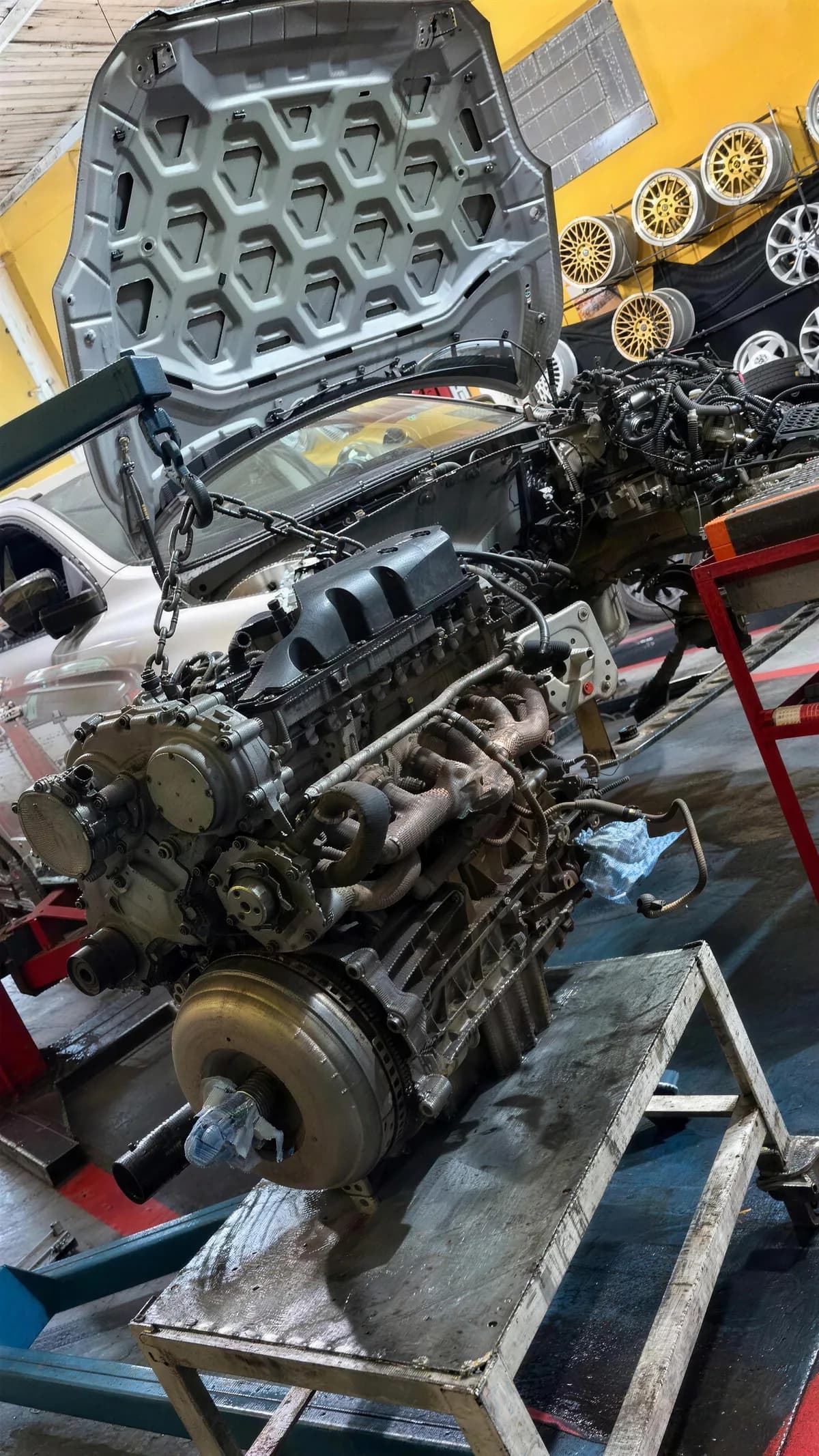 Motor de seis cilindros fora do carro, apoiado num cavalete, com o carro desmontado ao fundo